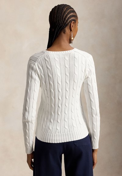 Sieviete ar sasietām matiem, valkājot baltu kabelknit džemperi un tumšas bikses, stāvot ar muguru pret kameru pret neitrālu fonu.