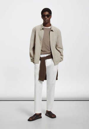 Homme portant une veste beige par-dessus une chemise marron, un pantalon blanc, un pull marron noué à la taille, des sandales foncées et des lunettes de soleil, debout devant un fond uni.
