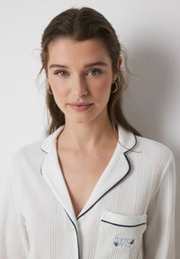 Weißes, geripptes Schlafanzug-Oberteil mit kontrastierendem, marineblauen Piping, einer aufgesetzten Tasche und besticktem Detail. Glatte Textur und klassisches Kragen-Design.