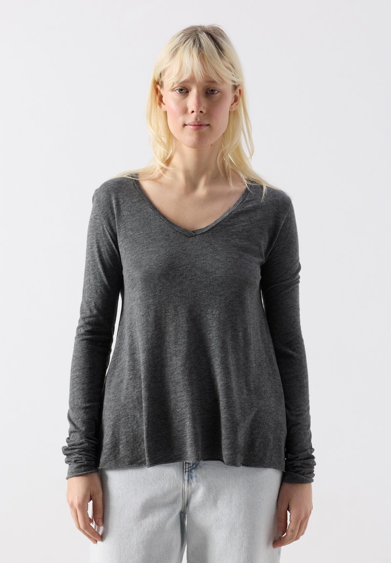 Gaiša blondīne, tērpusies tumši pelēkā garroku V-veida kakla topā un gaiši zilās džinsās, stāv pret vienkāršu baltu fonu.