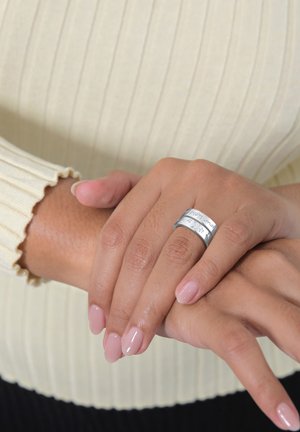 Anello d'argento con testo inciso, caratterizzato da una superficie liscia e lucida. Le mani sono appoggiate su un maglione cremoso e texturizzato.