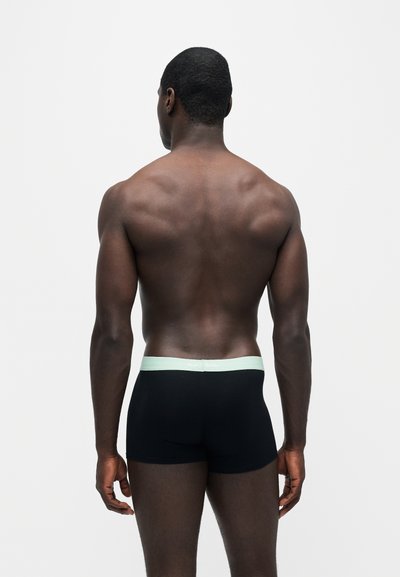 Homme musclé à la peau foncée, debout, tourné de dos, portant un boxer noir avec une ceinture claire sur un fond uni.