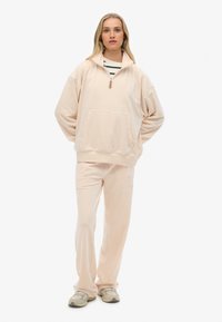 Pullover in pile rosa chiaro con mezza zip, tasca a marsupio e design ricamato; abbinato a pantaloni coordinati e sneakers beige.
