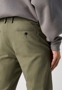 Khaki zelené kalhoty vyrobené z hladké tkaniny, s jednou zadní kapsou na knoflík a volným střihem, spárované se šedým topem s dlouhým rukávem.