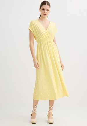 Mujer con un vestido midi amarillo claro con mangas cortas, escote en V, cintura fruncida y zapatos de cuña tipo espadrilles beige con tiras en el tobillo.