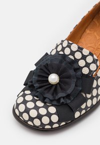 Chaussure plate à pois noirs et blancs avec fleur en tissu froncé noire et décoration centrale en perle sur le bout.