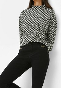 Blusa bianco e nero con colletto alto e motivo geometrico, abbinata a jeans neri con dettagli dorati e tasche laterali.
