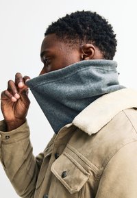 Szürke polár nyakmelegítő, amelyet egy világosbarna dzsekihez viselnek, amely krémszínű, texturált gallérral rendelkezik. A nyakmelegítő részben fel van húzva, hogy eltakarja az arcot.