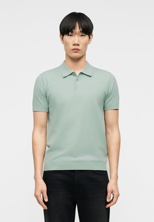 Junger Mann mit kurzen schwarzen Haaren, der ein hellgrünes, kurzärmeliges Poloshirt und eine dunkle Hose trägt, stehend vor einem schlichten weißen Hintergrund.