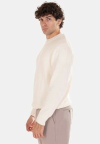 Maglione bianco lavorato a maglia con scollo rotondo, tessuto testurizzato e polsini a coste, abbinato a pantaloni beige chiaro. Design semplice e neutro.