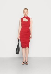 Vestido midi vermelho franzido com gola alta e detalhe recortado, confeccionado em tecido elástico. Acompanhado de uma bolsa preta e sandálias pretas com tiras.