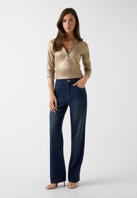 Gouden geribde top met lange mouwen en knopen, gecombineerd met wijde donkerblauwe denim jeans. Gouden sandalen met bandjes maken de outfit compleet.