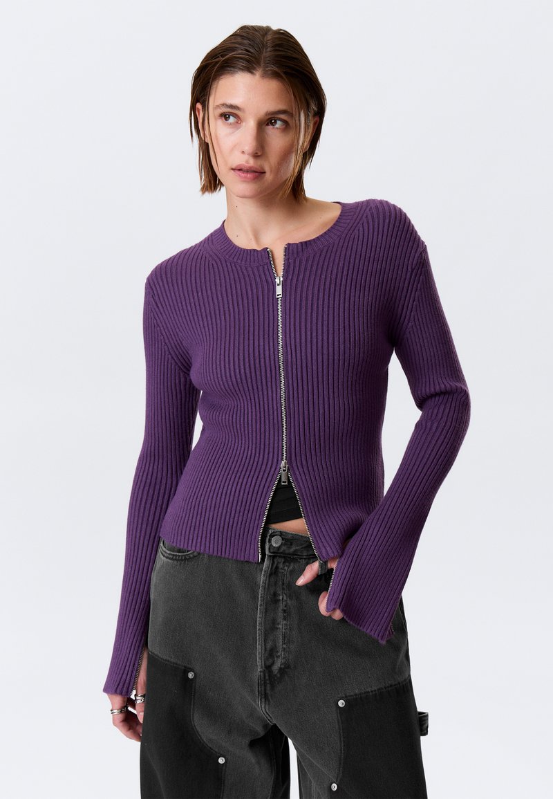 Cardigan violet en maille côtelée avec fermeture éclair à l'avant, manches longues et coupe ajustée. Orné d'un col montant et de poignets ouverts.