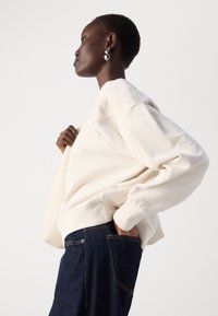 Felpa crema con design floreale testurizzato sulle spalle, caratterizzata da una vestibilità comoda e polsini a coste, abbinata a jeans in denim scuro.