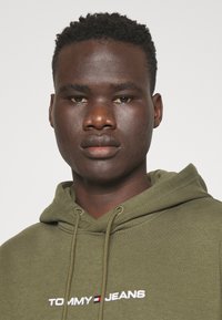 Sudadera verde oliva de tela suave, con capucha ajustable y un logotipo blanco "TOMMY JEANS" con acentos en rojo y azul.