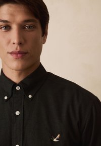 Čierna košeľa na gombíky z mäkkej tkaniny, s bielymi gombíkmi a farebne vyšitým vtákom na ľavej strane hrude.