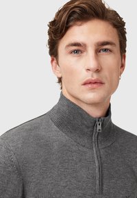 Grijze trui met ritssluiting en hoge kraag, gemaakt van zachte stof. Voorzien van een rits met een klein metalen trekhaakje en een geribbelde textuur bij de kraag.