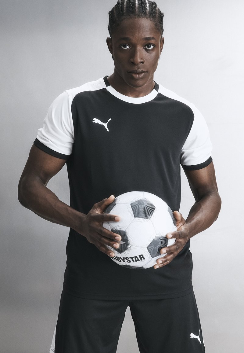 Athlet mit geflochtenem Haar, der schwarz-weiße Puma-Sportbekleidung trägt, hält einen schwarz-weißen Fußball auf Taillenhöhe vor einem grauen Hintergrund.