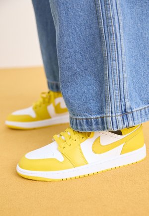 Zapatillas amarillas y blancas usadas con pantalones vaqueros azules, de pie sobre un suelo amarillo.