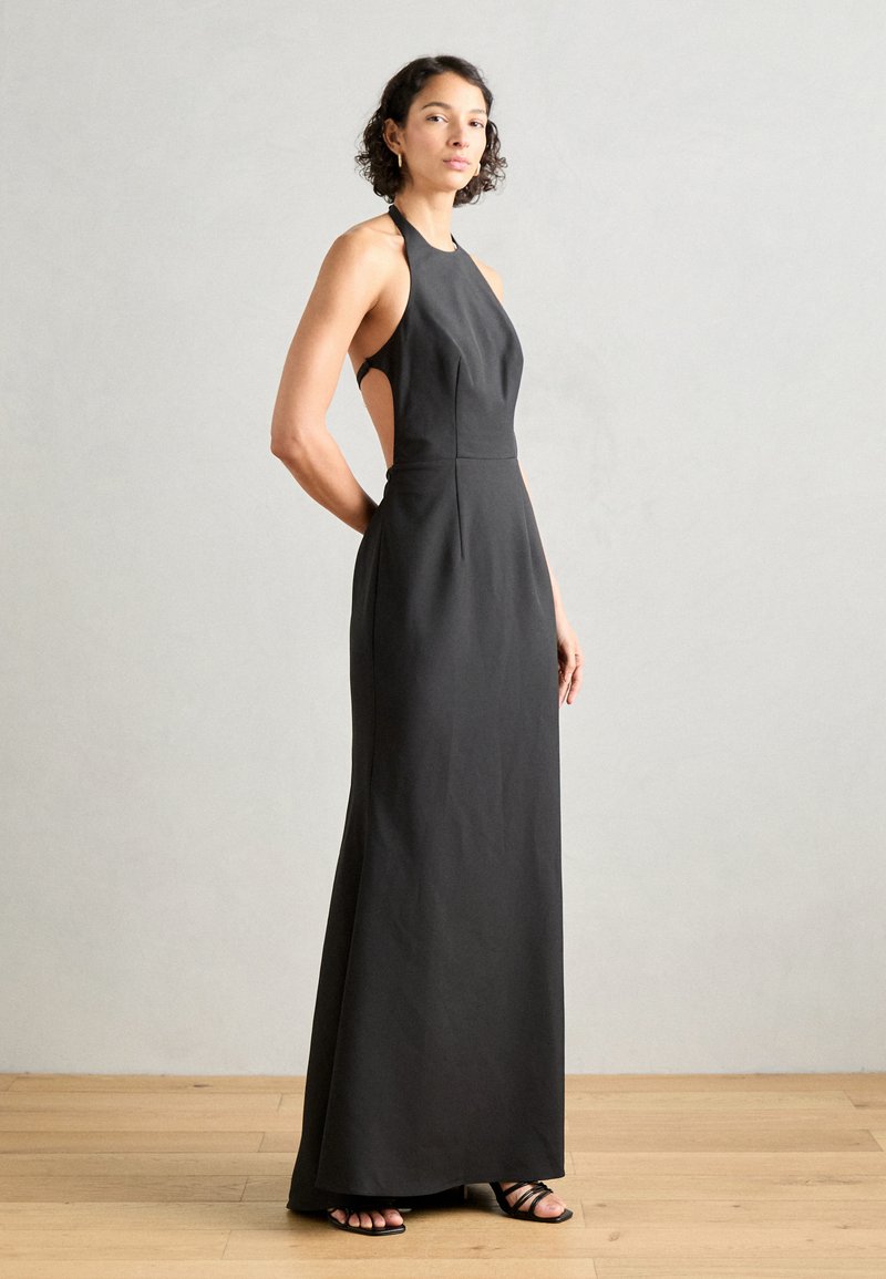 Črna halter obleka z izrezanimi stranicami, oprijetim zgornjim delom in padajočo krilo. Elegantna tkanina z minimalnimi šivi, v kombinaciji z črnimi sandali s trakovi.