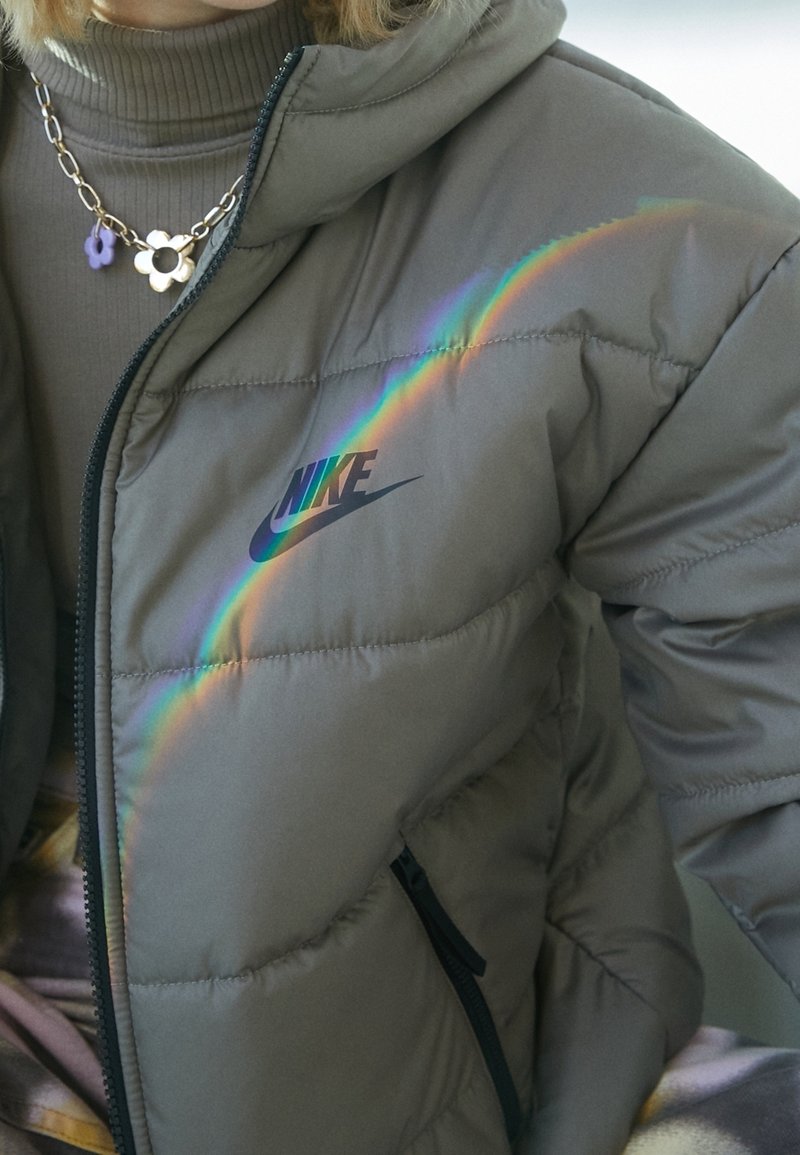 Olivengrüne Nylon-Pufferjacke mit schwarzem Reißverschluss, ausgestattet mit einem Nike-Logo und einem schimmernden Regenbogen-Effekt über der Brust. Sichtbare, strukturierte Steppung.