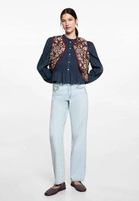 Broderad vinröd väst över en mörkblå blus, parat med ljusblå raka jeans och röda vävda flats. Strukturerad tygkontrast.