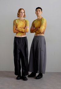 To modeller står side om side. Den venstre har på seg en langermet, tettsittende topp med et mønster i gult og brunt og cropped hem. Den høyre modellen har på seg løse, grå vidde bukser og svarte støvller.