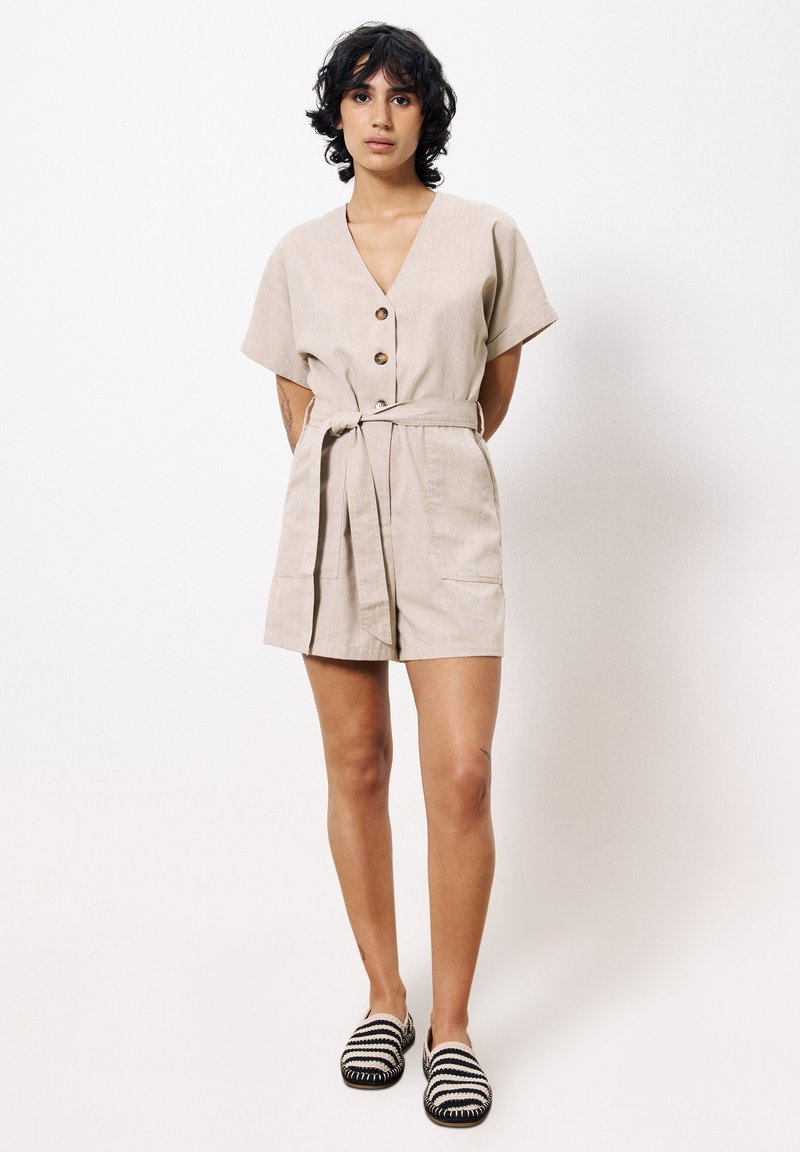 Jumpsuit beige court avec des manches courtes, décolleté en V, boutons en bois, ceinture à la taille et deux poches avant. Chaussures slip-on rayées noir et blanc.