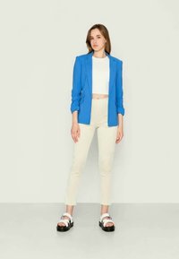 Blazer azul claro sobre um top cropped branco, combinado com calças cream de canalé e sandálias de plataforma pretas com tiras brancas. Texturas suaves.