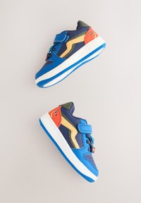 Baskets pour enfants en bleu, marine, vert et orange, dotées d'une sangle en velcro, de semelles en caoutchouc et d'un accent souriant sur le talon.