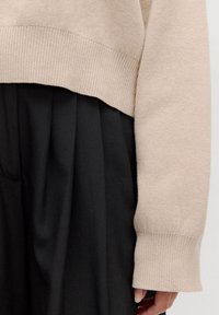 Beige, kurz geschnittener Pullover mit gerippten Bündchen, kombiniert mit einem schwarzen, plissierten Rock, der eine weiche Textur und glatte Stoffe zeigt.