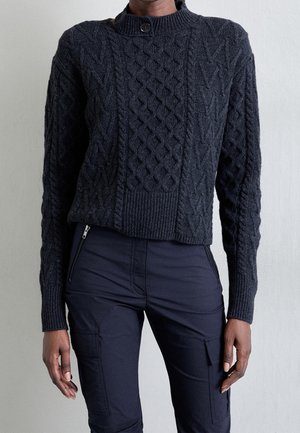 Personne portant un pull torsadé gris foncé avec un col boutonné et un pantalon cargo noir avec des poches zippées.