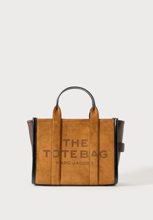 Sac fourre-tout en suédine marron avec des accents en cuir noir, doté de deux courtes poignées et du texte "LE SAC FOURRE-TOUT" en gras sur le devant.