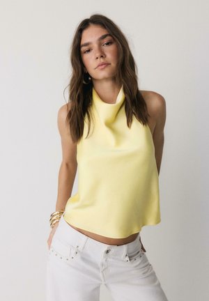 Mladá žena s dlhými hnedými vlasmi, oblečená v žltom tielku bez rukávov a bielych džínsoch, stojaca pred svetlým jednoduchým pozadím.