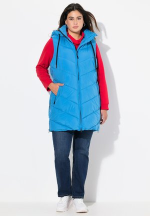 Blauer Puffweste mit Kapuze, ausgestattet mit einem gesteppten Muster und schwarzen Reißverschlussdetails. Getragen über einem roten Sweatshirt und kombiniert mit dunklen Jeans.