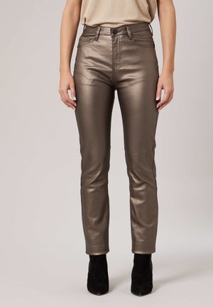 Pantalon slim en bronze métallique porté par une personne debout, bras détendus, associé à des bottines noires et un haut beige.