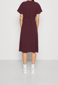 Femme debout, dos à nous, vêtue d'une robe violette mi-longue, de chaussettes blanches et de baskets épaisses blanches sur un fond clair uni.