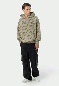 Hoodie verde oliva com padrão camuflado, com um bolso frontal e capuz ajustável, combinado com calças de carga pretas com múltiplos bolsos e ténis brancos.