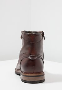 Brązowy skórzany botek za kostkę z okrągłym noskiem, bocznym zamkiem błyskawicznym i wzmocnioną tkaniną w pasiaki. Posiada delikatne przeszycia i drewnianą podeszwę.