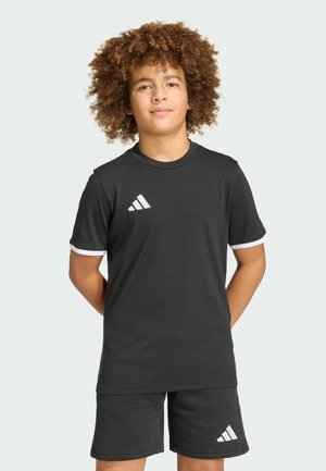 Junge mit lockigem Haar steht mit Händen hinter dem Rücken, trägt ein schwarzes Adidas-T-Shirt und Shorts mit weißen Logoakzenten.