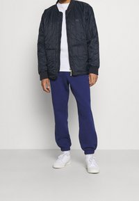 Svart quiltad bomberjacka med dragkedja framtill, grå sweatshirt och marinblå joggers. Vita sneakers fulländar outfiten.