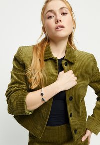 Chaqueta de pana verde oliva con textura acanalada, cuello redondo y botones negros. El modelo lleva una camiseta negra ajustada y una pulsera plateada con detalles oscuros.