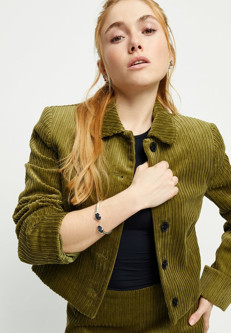 Chaqueta de pana verde oliva con textura acanalada, cuello redondo y botones negros. El modelo lleva una camiseta negra ajustada y una pulsera plateada con detalles oscuros.