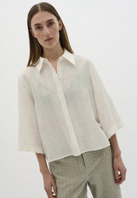 Camisa blanca texturizada con cuello abotonado, de corte holgado y mangas de tres cuartos, combinada con pantalones verdes claros con estampado.