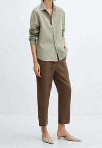 Chemise boutonnée vert olive avec une poche avant et des manches retroussées, associée à un pantalon marron fuselé et des mules beige clair. Design minimaliste.