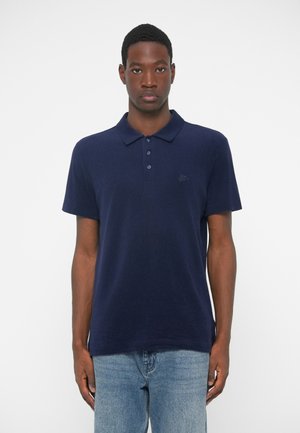 T-paita laivastonsinisestä pehmeästä kankaasta, jossa on napillinen kaulus, lyhyet hihat ja pieni brodeerattu logo rinnassa.