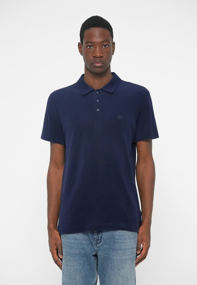 Tumši zilā polo krekls, kas izgatavots no mīksta auduma, ar pogu apkakli, īsām piedurknēm un mazu izšūtu logo uz krūšu.