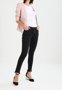 Jasnoróżowa marynarka na białej koszulce z czerwonym napisem, w połączeniu z czarnymi dżinsami z postrzępionym dołem i czarnymi szpilkami w kształcie migdałów.