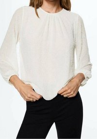 Blusa bianca texurizzata con maniche lunghe, scollatura arricciata e orlo leggermente curvo, indossata con pantaloni neri.