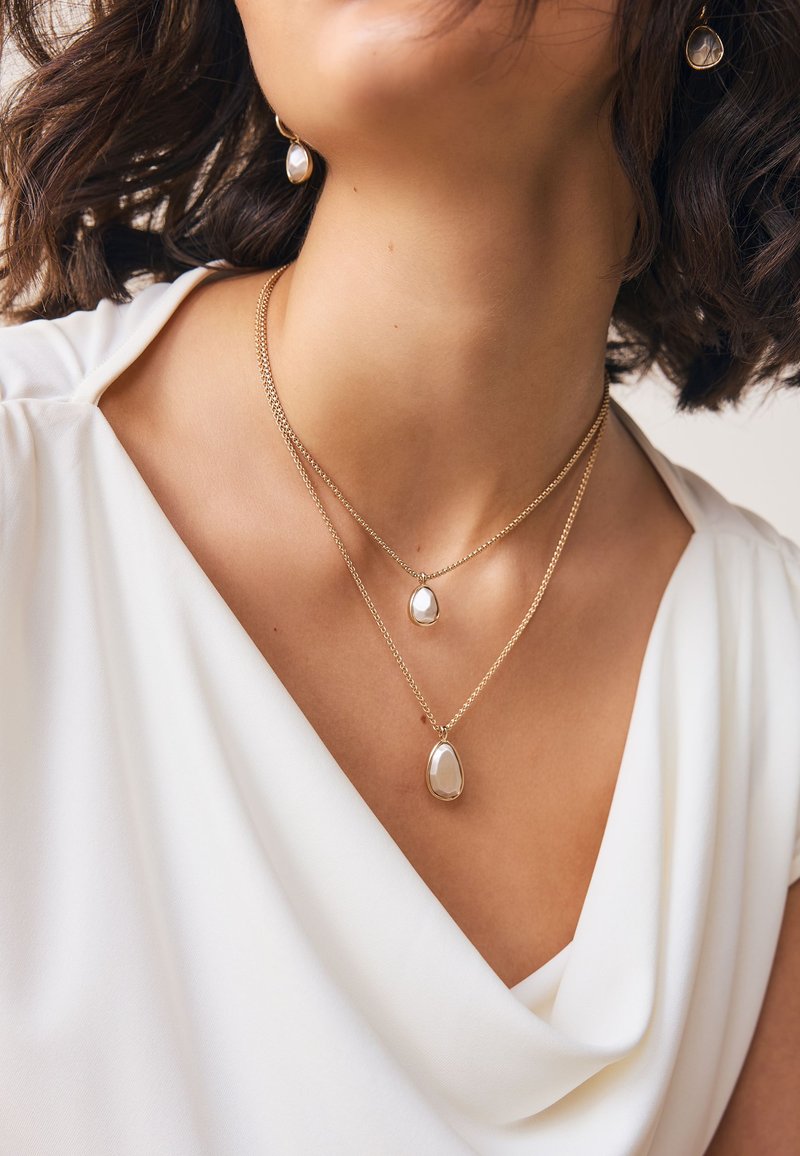 Woman wearing white blouse with layered gold necklaces and matching gold drop pearl earrings, showcasing jewelry details.